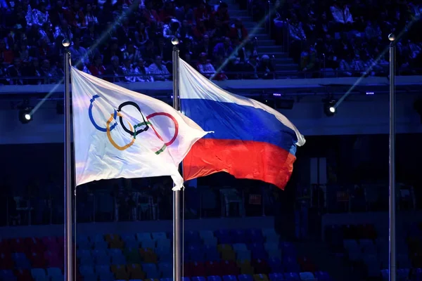 Drapeau olympique et drapeau russe sur des mâts lors d'une cérémonie dans un stade.