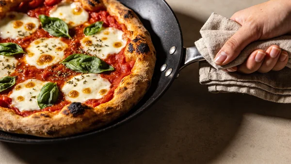 Pizza ronde dans une poêle antiadhésive noire, garnie de tomates, fromage fondu et herbes, vue de dessus avec main tenant la poignée de la poêle, bord croustillant visible, surface texturée