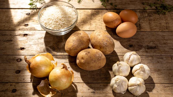 Gros plan en vue de dessus des cinq ingrédients essentiels disposés en cercle sur une table en bois : pommes de terre terreuse, riz blanc dans un bol en verre, trois œufs bruns, deux oignons dorés, et plusieurs gousses d'ail, éclairage chaud naturel