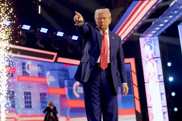 Donald Trump en costume bleu et cravate rayée rouge pointant vers l'avant sur une scène.