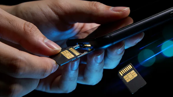 Mains tenant deux cartes SIM, l'une étant retirée d'un smartphone, l'autre insérée, visualisation du concept de SIM-swapping, lumière bleutée de l'écran du téléphone, arrière-plan sombre avec reflets technologiques