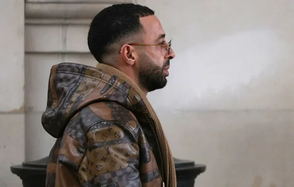 Le rappeur Naps portant une veste camouflage et une écharpe marron.