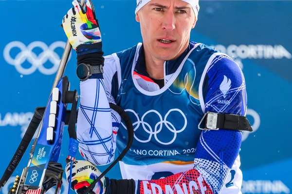 Un biathlète français masculin en plein effort sur une piste de neige, portant son fusil sur le dos, le visage concentré et déterminé, le paysage montagneux en arrière-plan