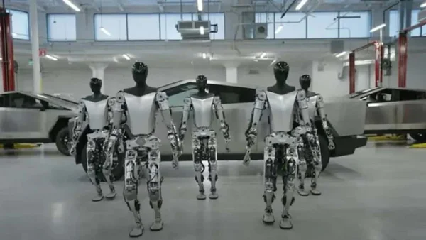 Cinq robots humanoïdes Tesla alignés devant une voiture dans un hall industriel.