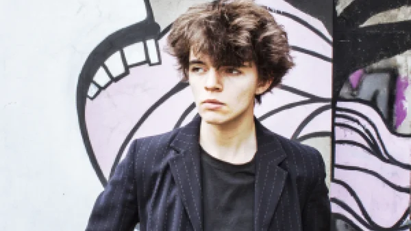 Sam Sauvage pose devant un mur urbain tagué, vêtu de son inévitable blazer rayé noir.