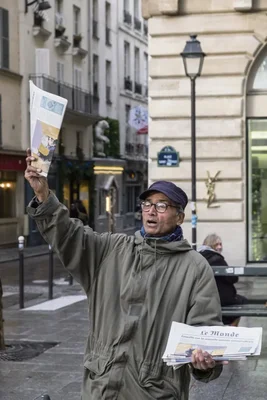 Un vendeur de journaux dans la rue, en veste verte et casquette bleue, levant un exemplaire du Monde avec une pile d'autres journaux.