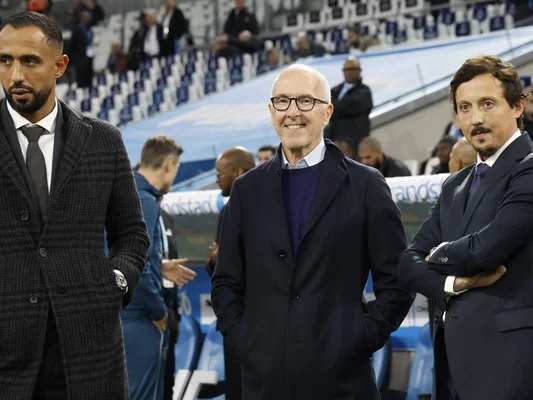 Trois hommes en tenue formelle sur une ligne de touche de stade, entourés de spectateurs.
