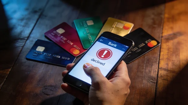 Vue en gros plan d'une main jeune tenant un smartphone affichant une application bancaire avec notification de refus, entouré de plusieurs cartes bancaires colorées éparpillées sur une table en bois, éclairage dramatique avec ombres, ambiance tendue