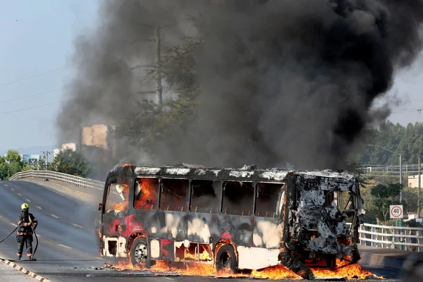 Un bus en feu sur une route, avec un pompier intervenant à proximité.
