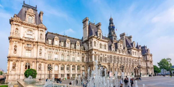 Façade ornée de l'Hôtel de Ville de Paris sous un ciel bleu avec une fontaine au premier plan.