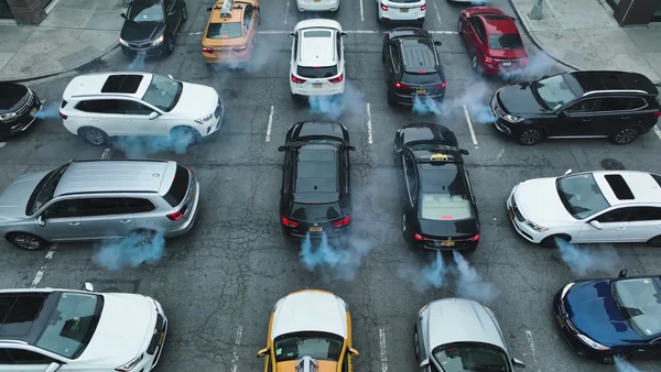 Vue en plongée verticale d'un embouteillage dense en centre-ville avec circulation aux heures de pointe, les fumées d'échappement bleutées s'échappent des pots arrière et flottent au-dessus de la route bitumée grise sous un ciel grisâtre