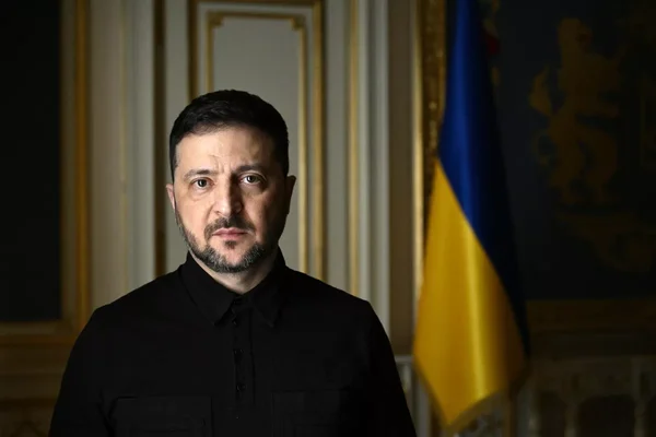 Volodymyr Zelensky, portant une chemise noire, se tient devant un drapeau ukrainien et des boiseries décoratives.