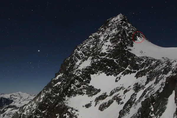 Un sommet montagneux enneigé sous un ciel étoilé, avec un cercle rouge indiquant une zone sur la crête.