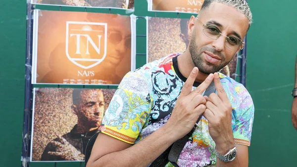 Le rappeur Naps portant une chemise colorée devant un mur vert.