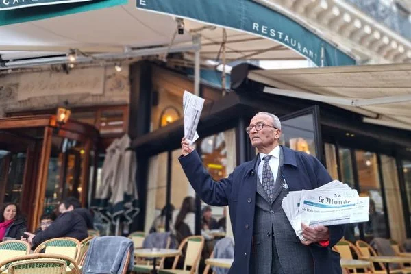 Ali Akbar, en manteau foncé et cravate, se tient devant un restaurant avec une pile de journaux Le Monde à la main.