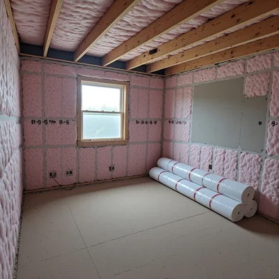 Vue de l'intérieur d'un appartement avec des travaux d'isolation en cours, des rouleaux de laine de verre posés contre un mur et une fenêtre ouverte laissant entrer la lumière