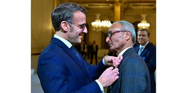 Une cérémonie formelle où un homme en costume bleu épingle une médaille sur le revers d'Ali Akbar, en costume gris, dans une salle ornée de lustres.