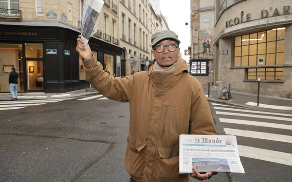 Un homme vend des journaux dans une rue de Paris, tenant un exemplaire en main, vêtu d'une veste beige et d'une casquette.