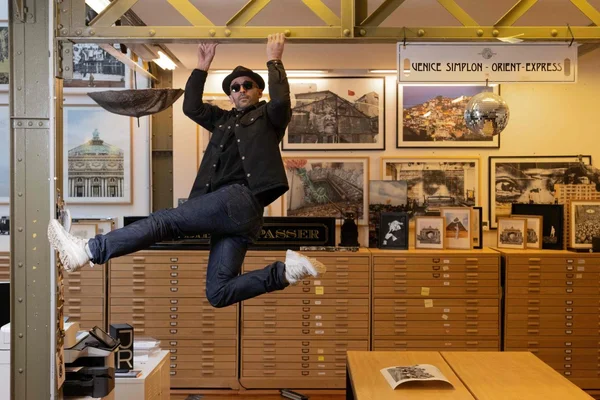 JR suspendu à une poutre métallique jaune dans une galerie d'art entouré de tableaux encadrés.