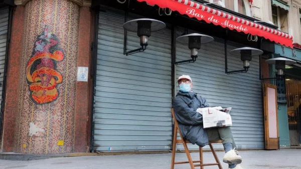 Un homme assis sur une chaise en bois devant un bar, portant un masque et tenant un journal, avec des rideaux métalliques fermés en arrière-plan.