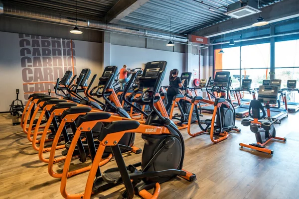 Salle de sport moderne avec équipements cardio orange et noir et deux personnes en train de faire de l'exercice.