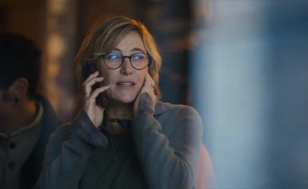 Une femme avec des lunettes et un pull gris-vert téléphone, main sur le visage.