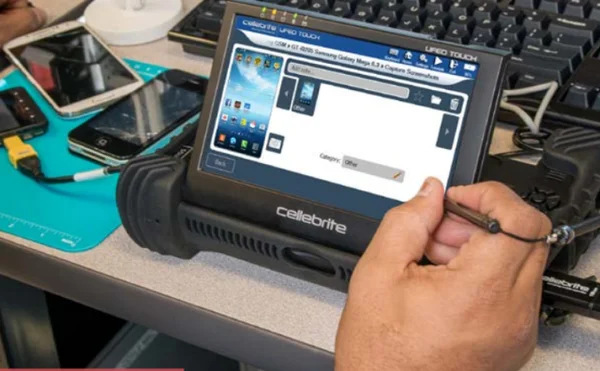 Une main utilise un stylet sur l'écran tactile d'un appareil Cellebrite posé sur un bureau avec des téléphones.