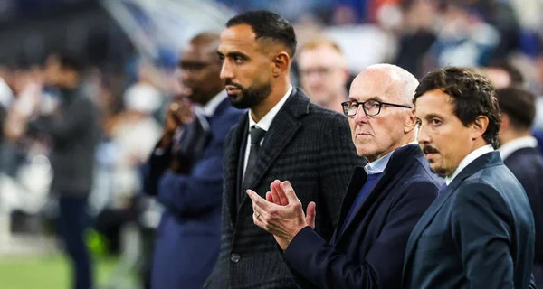 Quatre hommes en costumes sur un terrain de sport, l'homme du centre applaudissant.