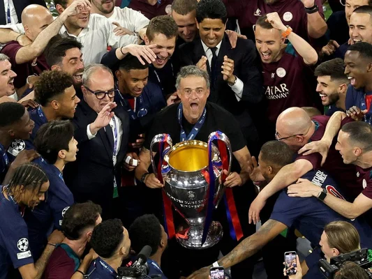 Un groupe de joueurs en tenue maillot marine et bordeaux célèbre en soulevant un trophée aux rubans rouges et bleus.