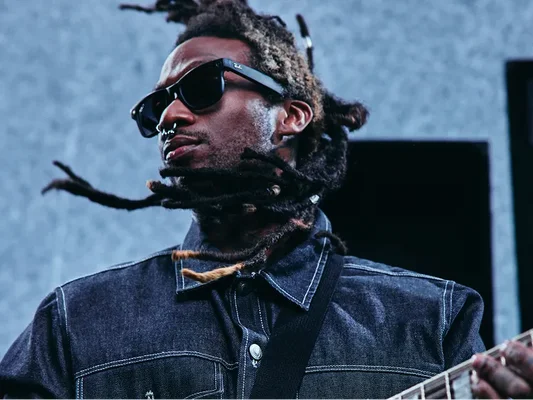 Une personne aux dreadlocks porte des lunettes et tient une guitare.