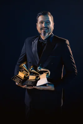 Un homme en costume sombre tient trois trophées Grammy devant un fond sombre.