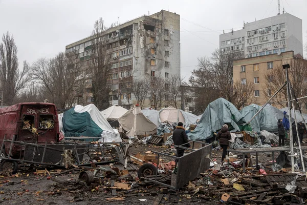Un quartier urbain en ruines avec des tentes de fortune et des débris jonchant le sol, présence d'une fourgonnette rouge.