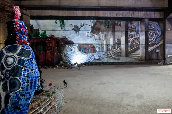 Fresque d'une ville en ruine végétalisée et sculpture colorée dans le parcours Les Extatiques.