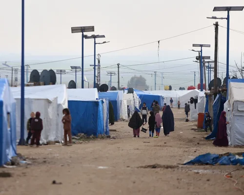 Plusieurs femmes en abayas noires et des enfants marchent le long d'une rangée de tentes.