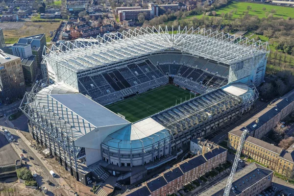 Vue aérienne d'un stade moderne présentant une toiture en treillis métallique et un terrain vert, situé au milieu d'une zone urbaine.
