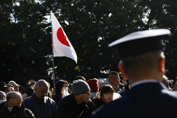 Une foule rassemblée à l'extérieur avec un drapeau japonais en arrière-plan et un homme en uniforme au premier plan.