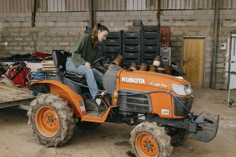 Agriculteur en vert et jeans assis sur son tracteur à l'intérieur d'une structure agricole.