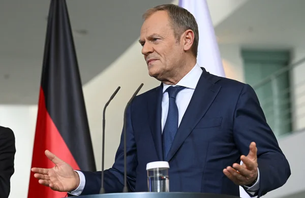 Donald Tusk, en costume bleu, parlant à un podium avec un drapeau allemand en arrière-plan.