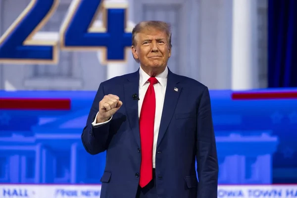 Donald Trump en costume bleu marine, le poing levé, lors d'un événement public avec le chiffre 24 en arrière-plan.