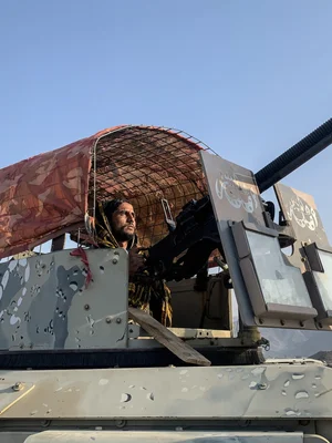 Un homme portant un couvre-chef jaune et noir actionne une mitrailleuse installée sur un véhicule militaire camouflé.