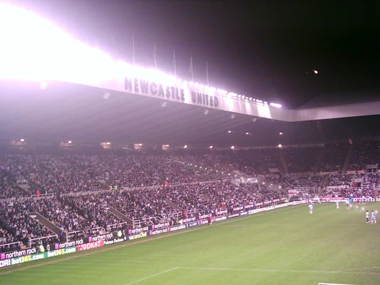 Intérieur du stade St James' Park de nuit avec le sigle lumineux NEWCASTLE UNITED dominant les tribunes lors d'une rencontre.