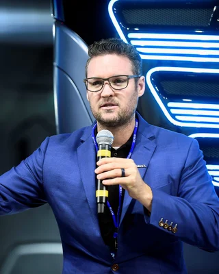 Trevor Milton tenant un micro devant un véhicule illuminé en bleu lors d'un événement.