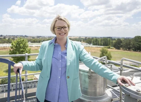 Une femme en veste bleu clair devant des équipements industriels avec un paysage vert en arrière-plan.