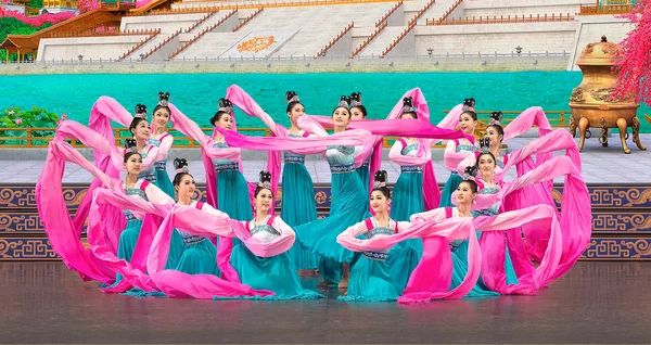 Un groupe de danseurs en costumes traditionnels bleus et jaunes performe sur scène avec des rubans roses.