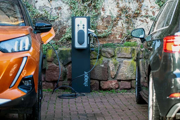 Une borne de recharge Hager entre deux voitures, l'une branchée, devant un mur en pierre végétalisé.