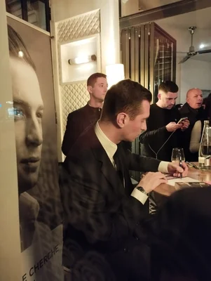 Jordan Bardella en séance de dédicace, assis à une table en compagnie d'autres personnes.