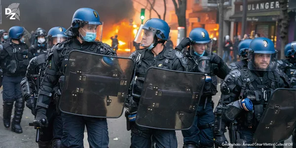 Une ligne de policiers anti-émeute avançant devant des incendies lors de troubles civils.