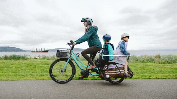 Une femme portant un casque conduit un vélo cargo turquoise avec deux enfants sur l'arrière, près d'un plan d'eau.