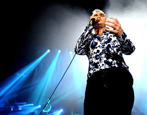 Morrissey sur scène, gestes expressifs, sous un éclairage bleu et une atmosphère enfumée.