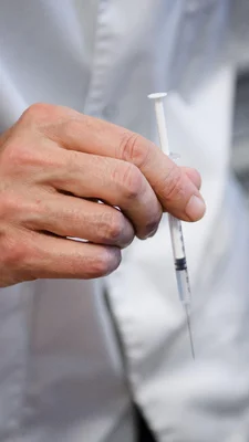 Une main vide pose une pilule sur une table de nuit en bois à côté d'un réveil matin, la lumière du matin filtre à travers les rideaux à demi fermés